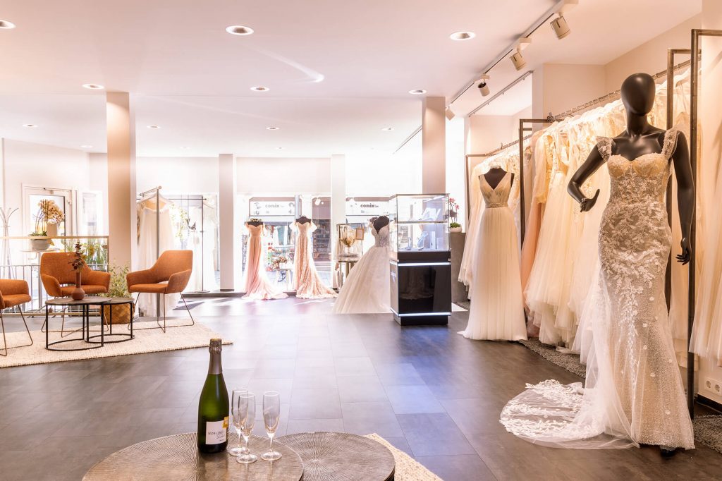 Brautkleider, elegant präsentiert in eleganter Atmosphäre in dem Bridal und Groom Store, einem Brautmodeladen in Lörrach. Im Vordergrund ist eine Sitzecke mit einem kleinen Tisch - gedeckt mit zwei Gläsern und einem kalten Sekt.