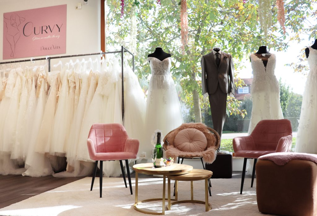 Ein stilvoll eingerichteter Brautmodenladen mit einem speziellen Curvy-Bereich, Dolce Vita Hügelheim-Müllheim. Zu sehen sind weiße Brautkleider auf Kleiderstangen, zwei ausgestellte Hochzeitskleider auf Puppen und ein eleganter Hochzeitsanzug. Der Raum ist lichtdurchflutet, mit großen Fenstern, dekorativen Blumenarrangements und gemütlichen rosa Sesseln für die Beratung. Ein Tisch mit Sektflasche und Gläsern rundet das Ambiente ab.